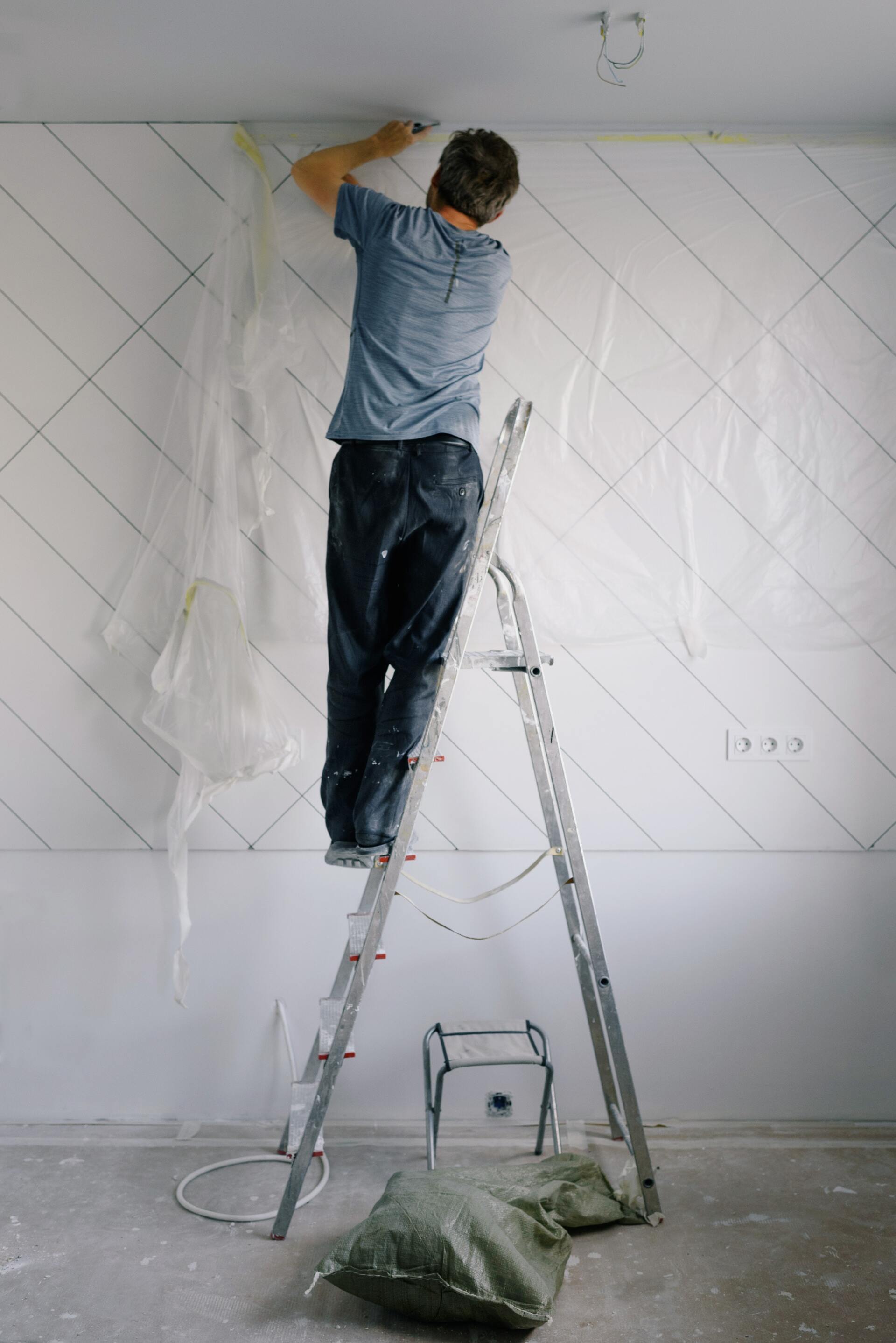 Person painting a white wall while standing on a ladder in a room under renovation