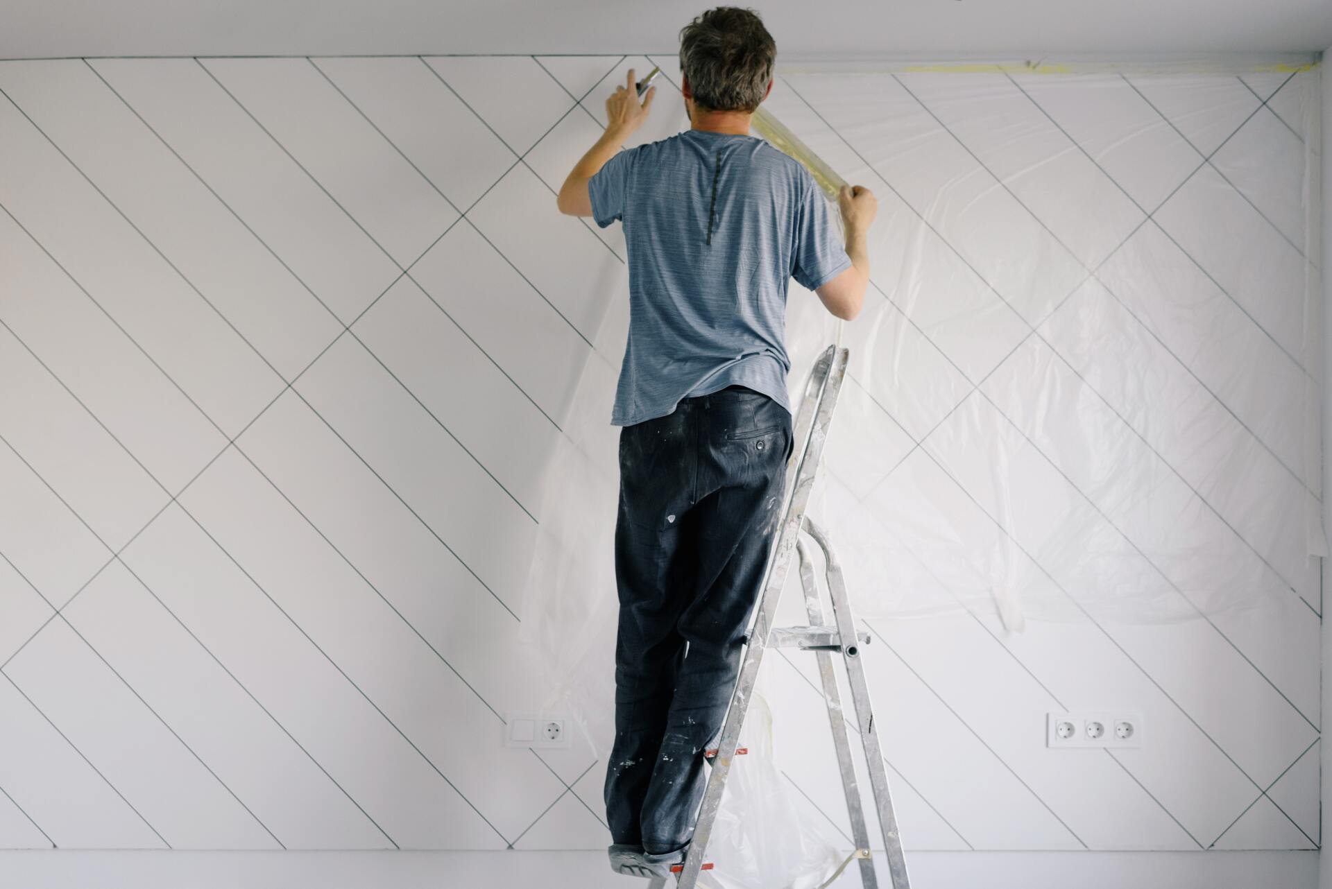 Person standing on a stepladder, painting diagonal lines on a white wall.