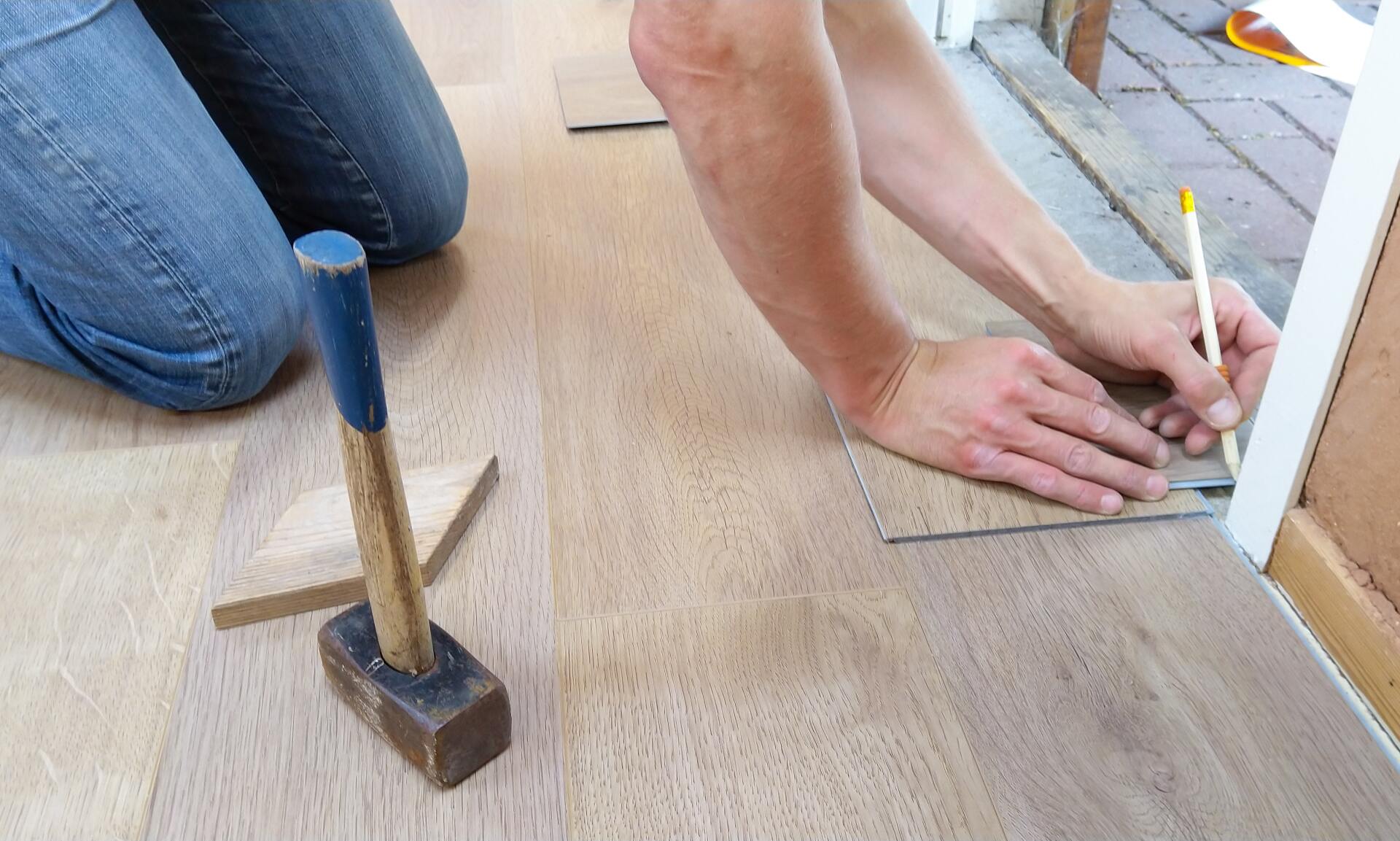 Person marking a wood floor seam with a pencil while another person kneels beside a hammer and chisel.