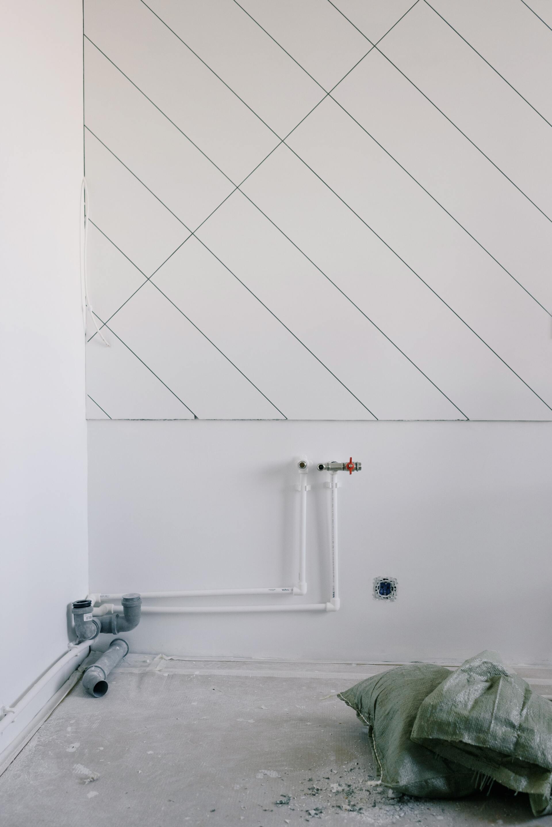Partially finished room wall with diagonal tile pattern, exposed pipes, and construction materials on the floor