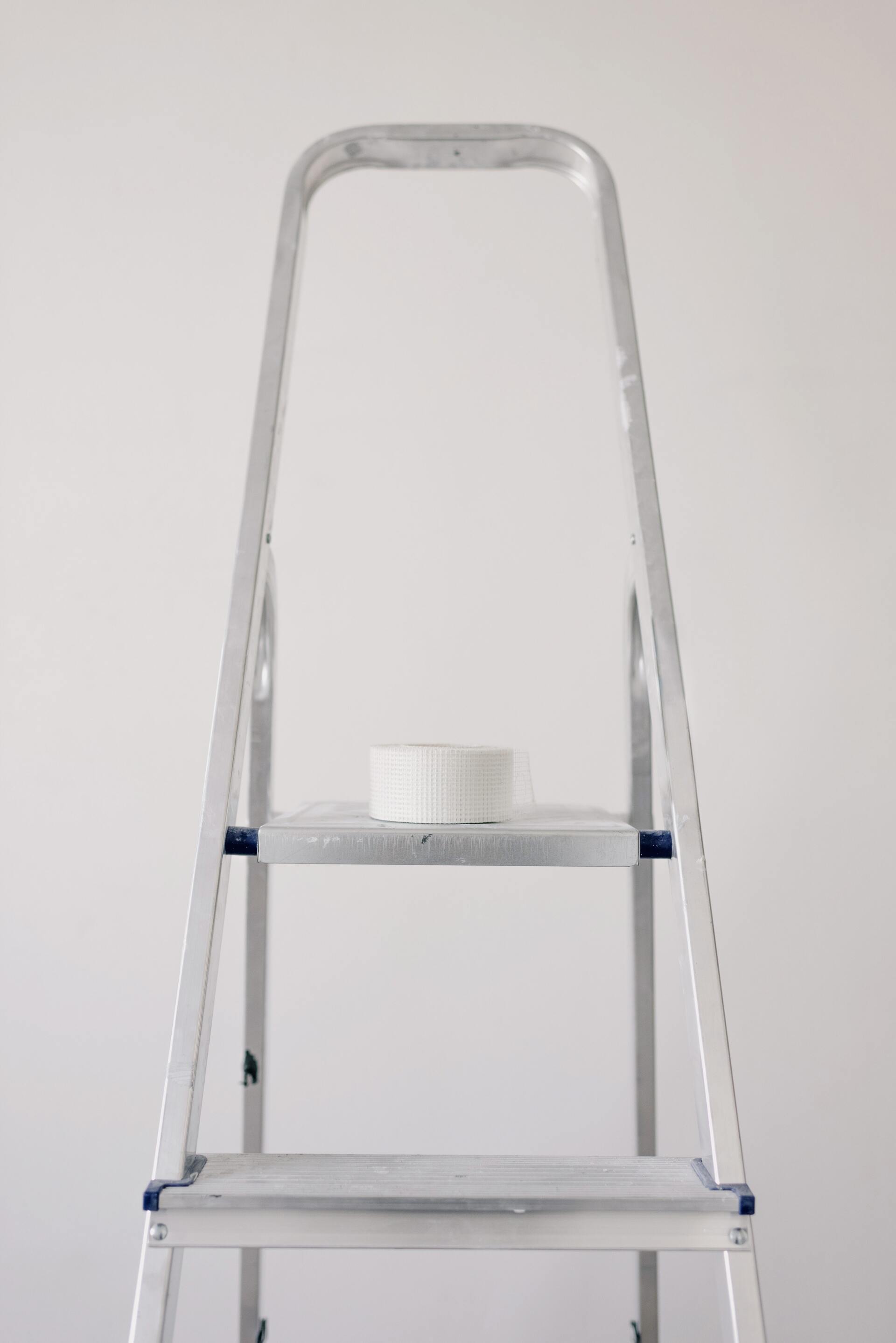 Silver step ladder with a white roll of tape on the top platform against a plain wall